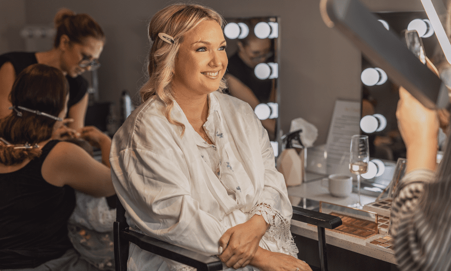 Bride smiling while getting her hair and makeup done, surrounded by beauty professionals in a well-lit dressing room on her wedding day.