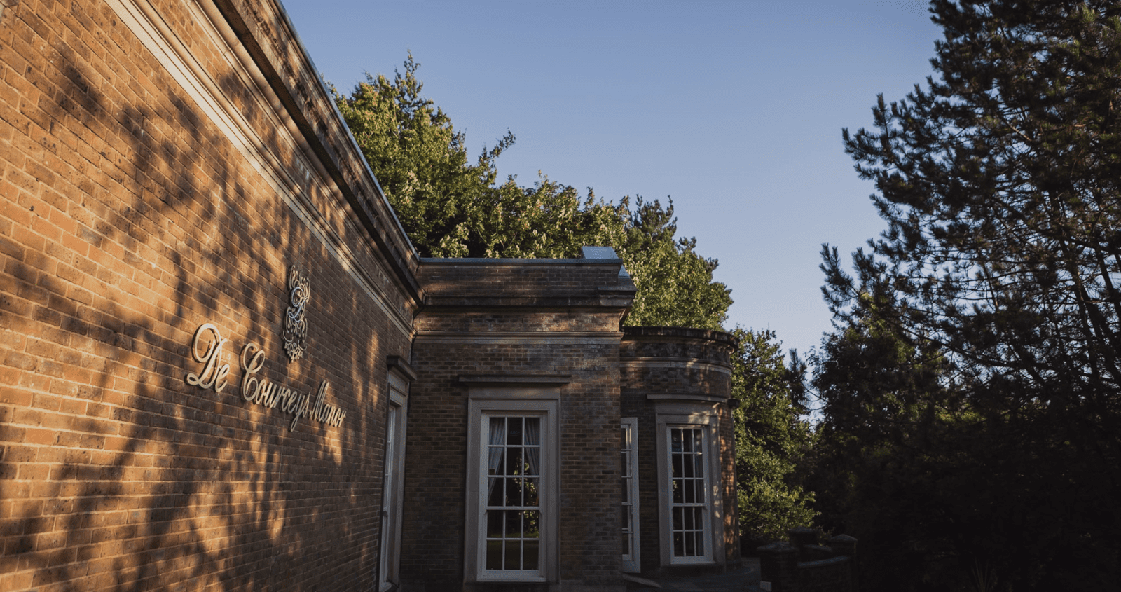 Exterior view of De Courceys Manor, a historic brick building surrounded by lush greenery under a clear blue sky in January