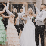 A joyful wedding celebration with a bride, groom, and guests popping champagne outside a brick manor venue, all dressed in formal attire, smiling and laughing.