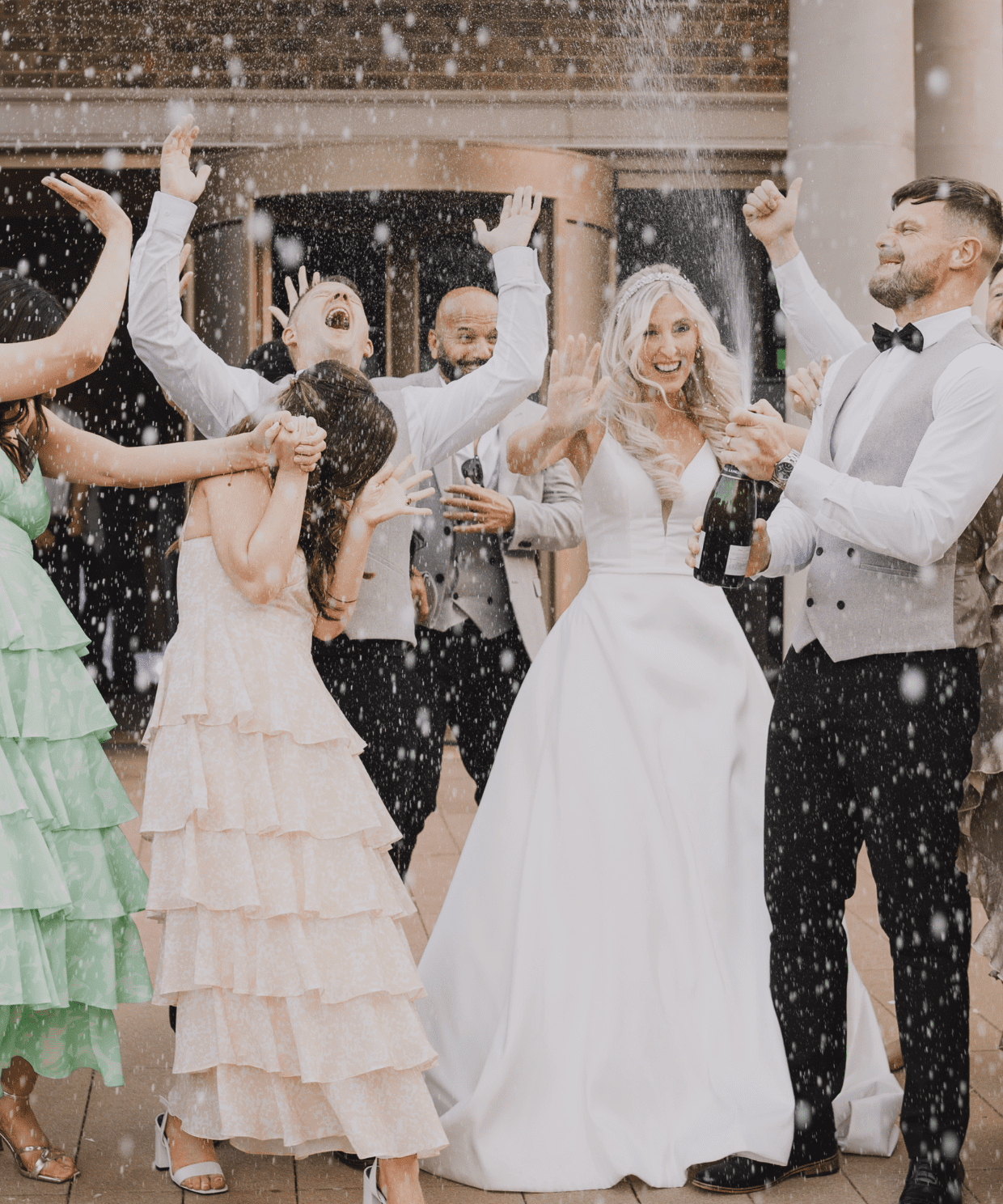 A joyful wedding celebration with a bride, groom, and guests popping champagne outside a brick manor venue, all dressed in formal attire, smiling and laughing.