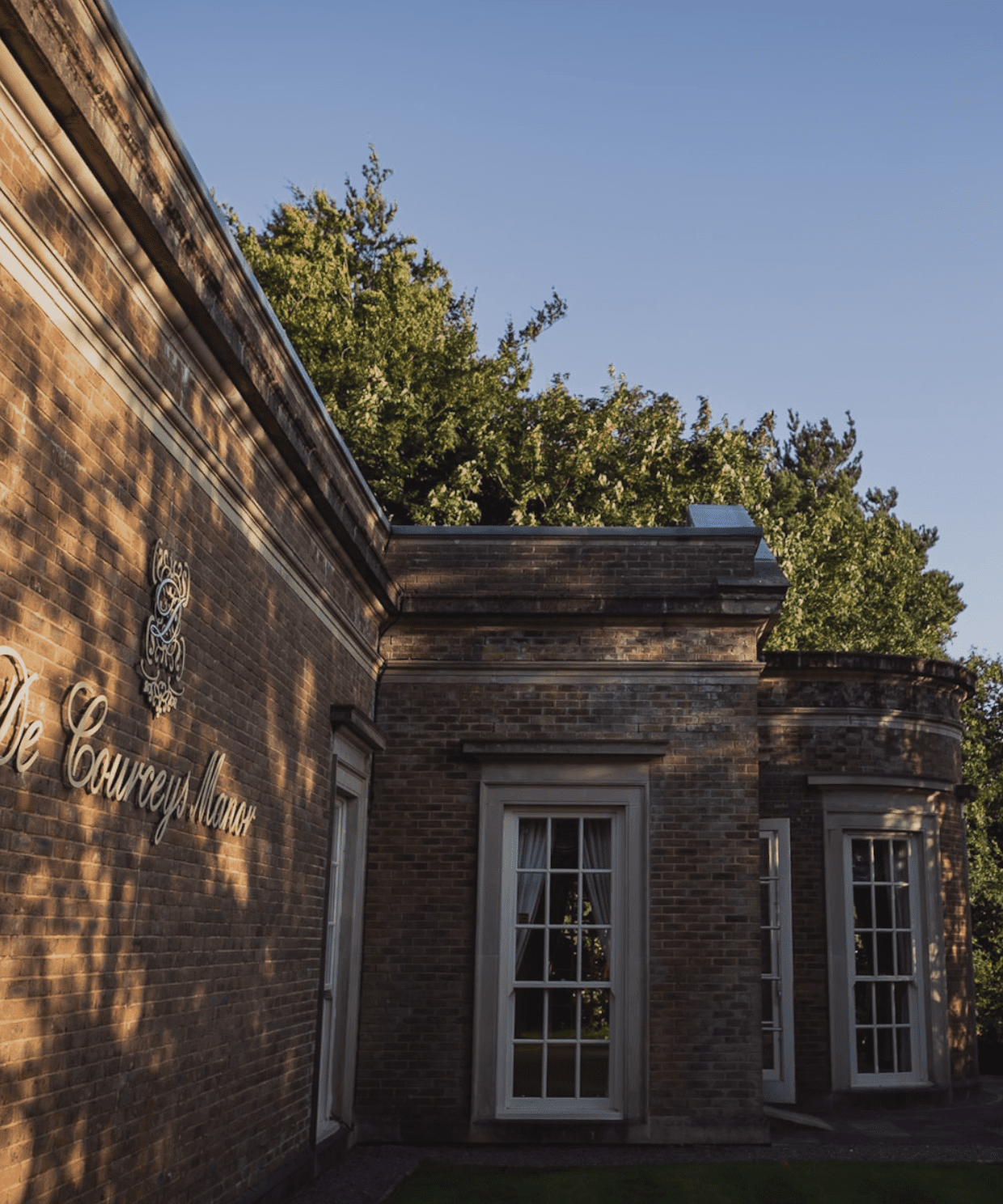 Exterior view of De Coucy Manor, a historic brick building surrounded by lush greenery under a clear blue sky.